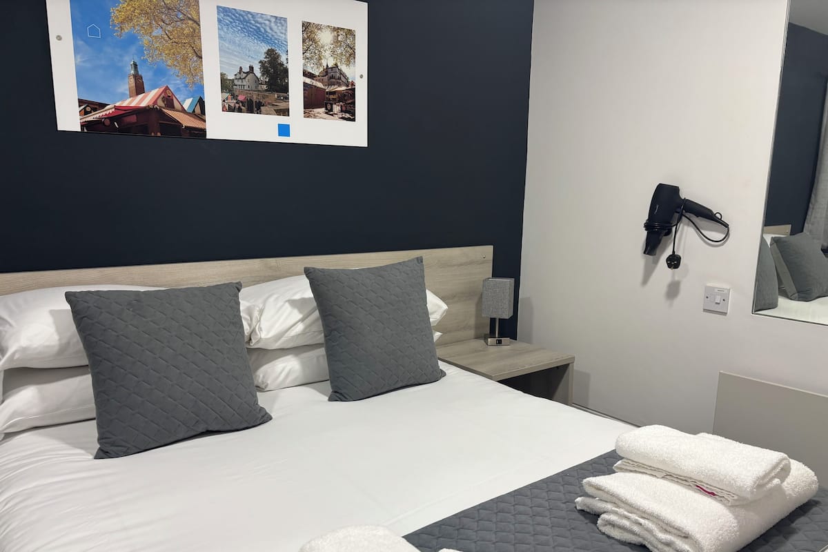 A neatly made bed with grey and white bedding, a bedside table with a lamp, and a wall-mounted hairdryer.