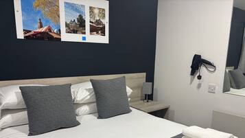 A neatly made bed with grey and white bedding, a bedside table with a lamp, and a wall-mounted hairdryer.