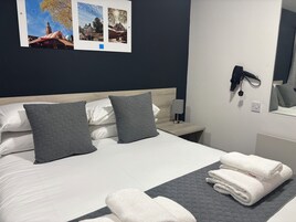 A neatly made bed with grey and white bedding, a bedside table with a lamp, and a wall-mounted hairdryer.