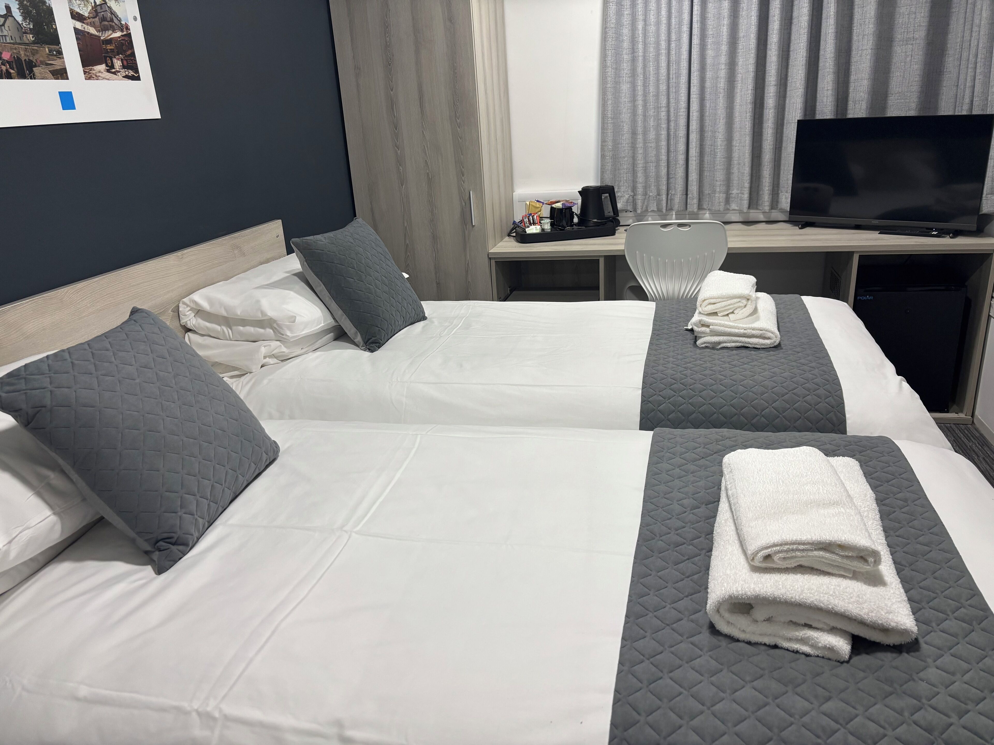 Ett hotellrum med två sängar, där varje säng har vita och grå sängkläder, en platt-TV, ett litet bord med en kaffebryggare och ett fönster med gardiner.