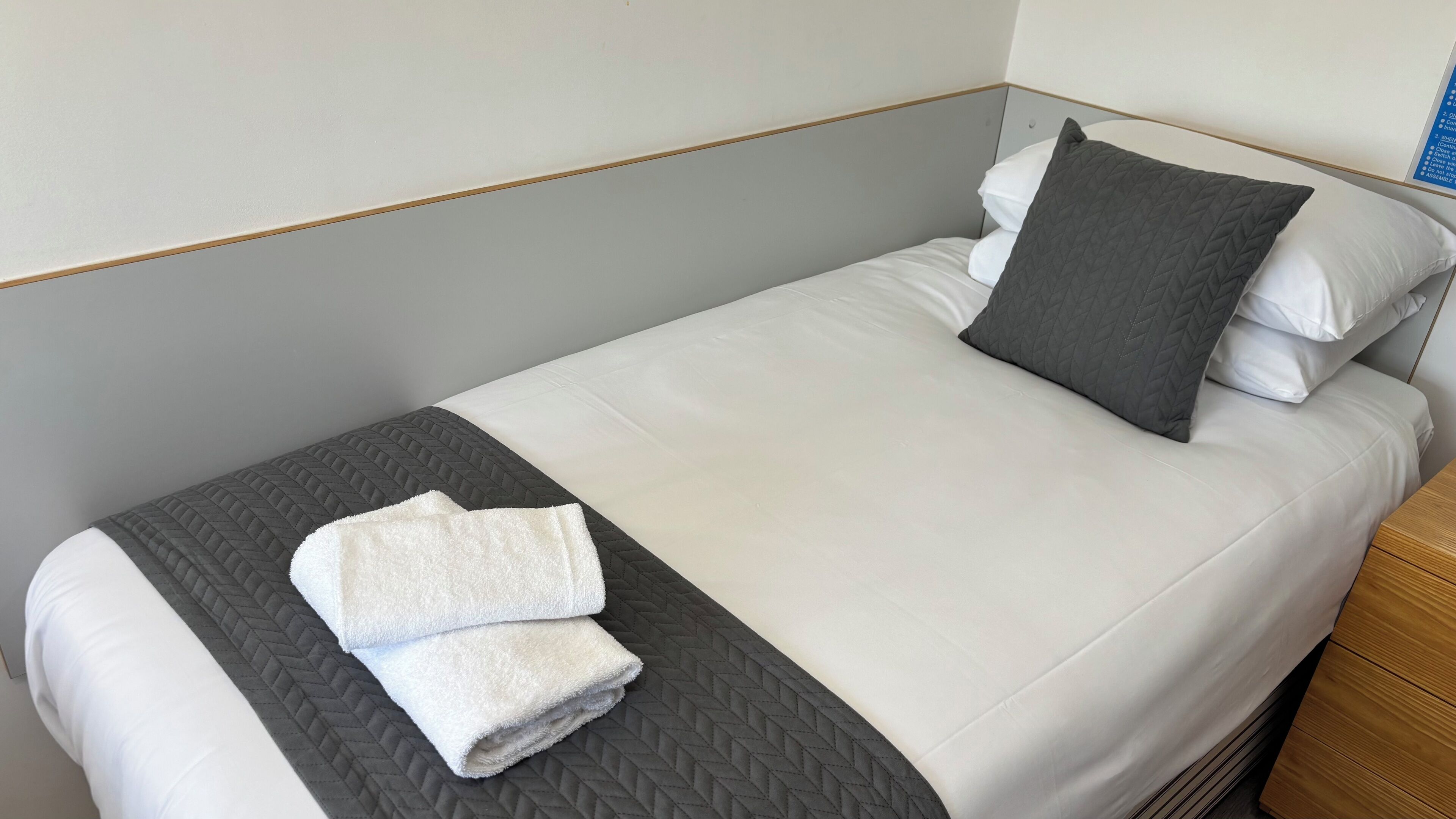 Una cama con ropa blanca bien tendida, una almohada acolchada gris y una toalla blanca doblada sobre una corrida de cama gris.
