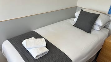 Una cama con ropa blanca bien tendida, una almohada acolchada gris y una toalla blanca doblada sobre una corrida de cama gris.