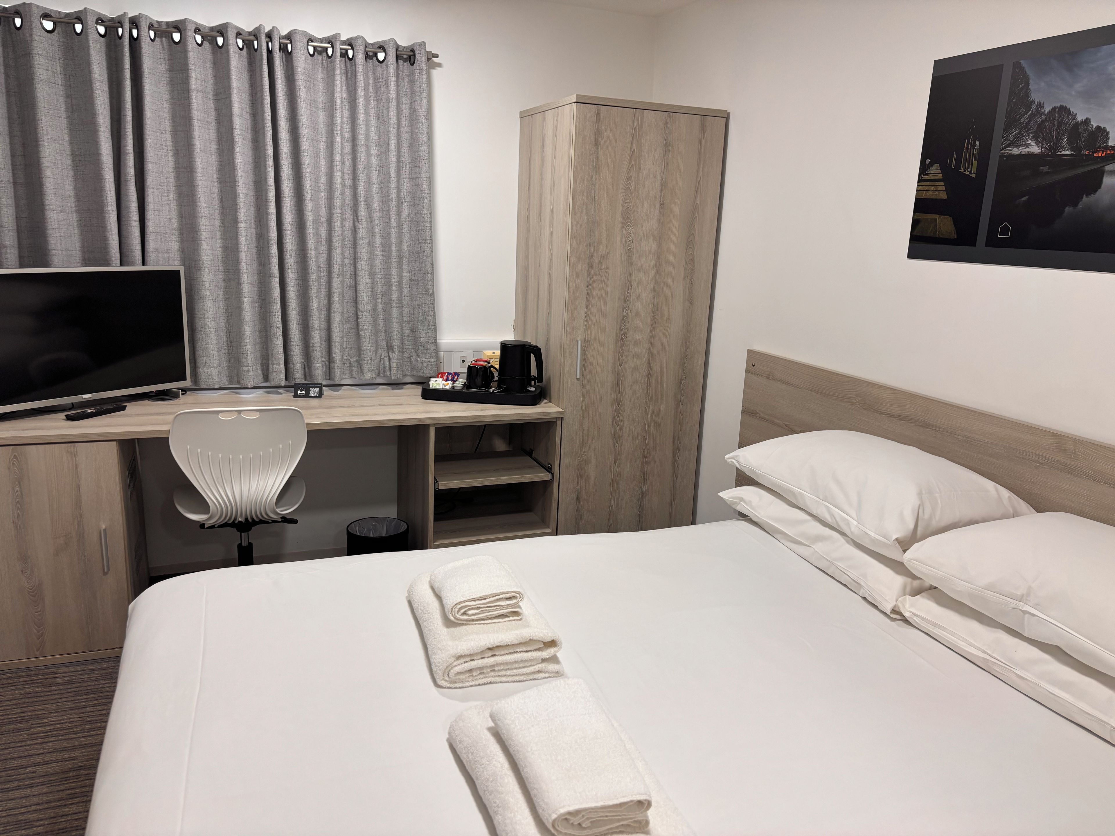 Habitación de hotel con cama, escritorio con computadora, silla, armario y un cuadro en la pared.