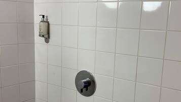 Un baño con paredes de azulejos blancos, una ducha con cabezal de ducha manual, un inodoro y un desagüe en el suelo.