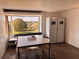 A hotel room with a large window, a dining table with chairs, a refrigerator, and a note on the door.