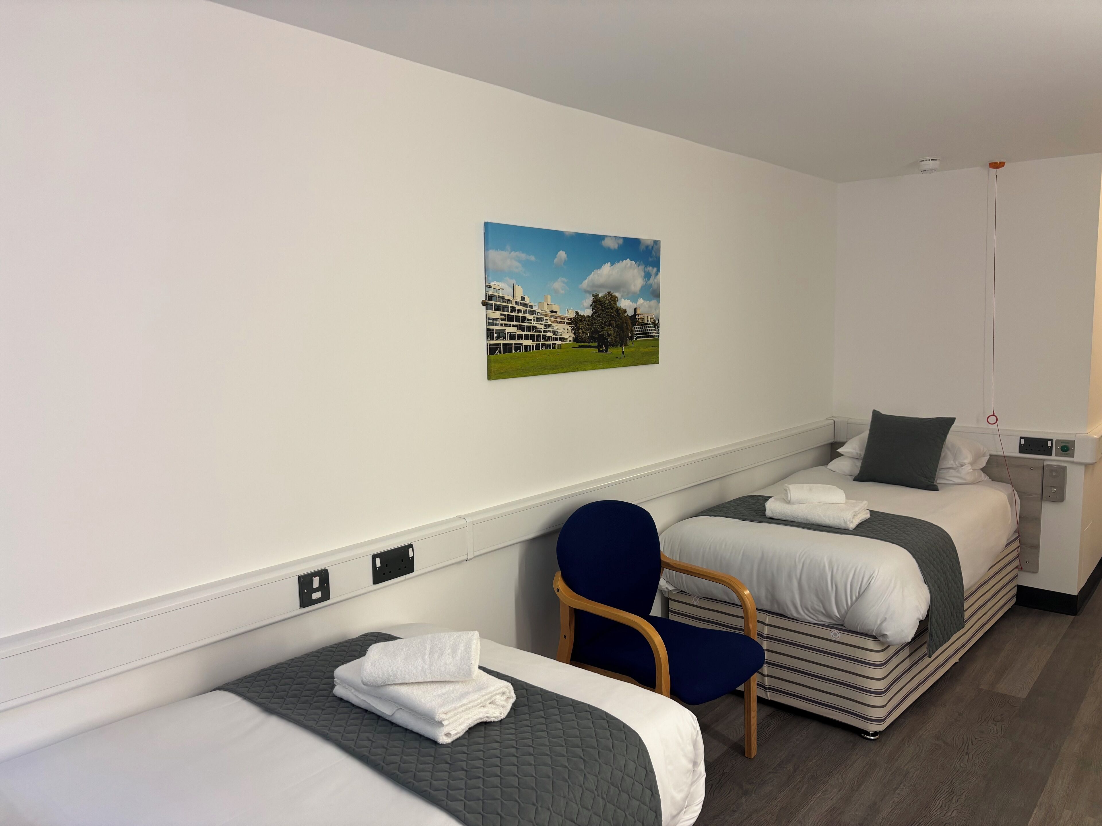 A hotel room with two single beds, a chair, a painting of a building and trees, and a small bedside table with a lamp.