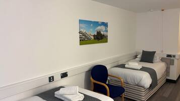 A hotel room with two single beds, a chair, a painting of a building and trees, and a small bedside table with a lamp.