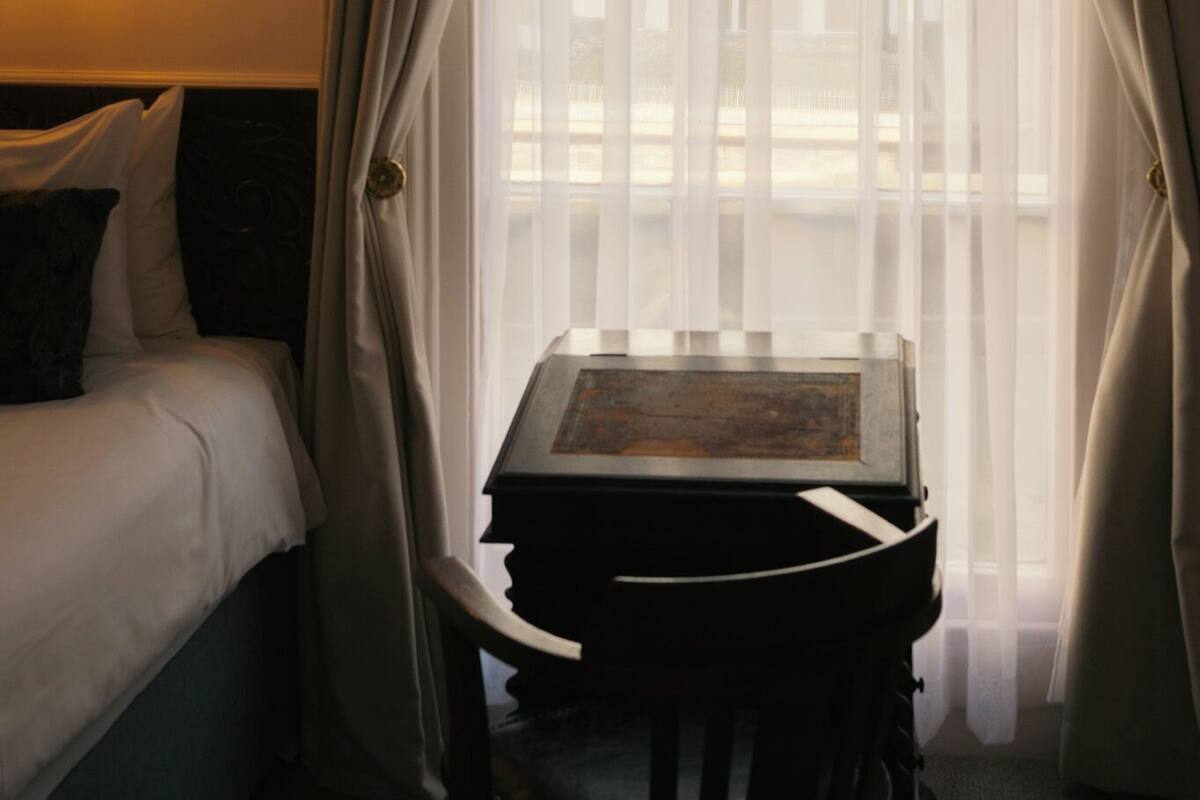 A hotel room with a bed, a table with a chair, and sheer curtains.