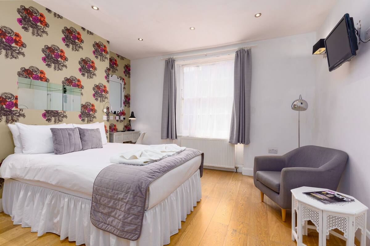 A bedroom with a bed, a chair, a small table, a mirror, and a window with curtains.