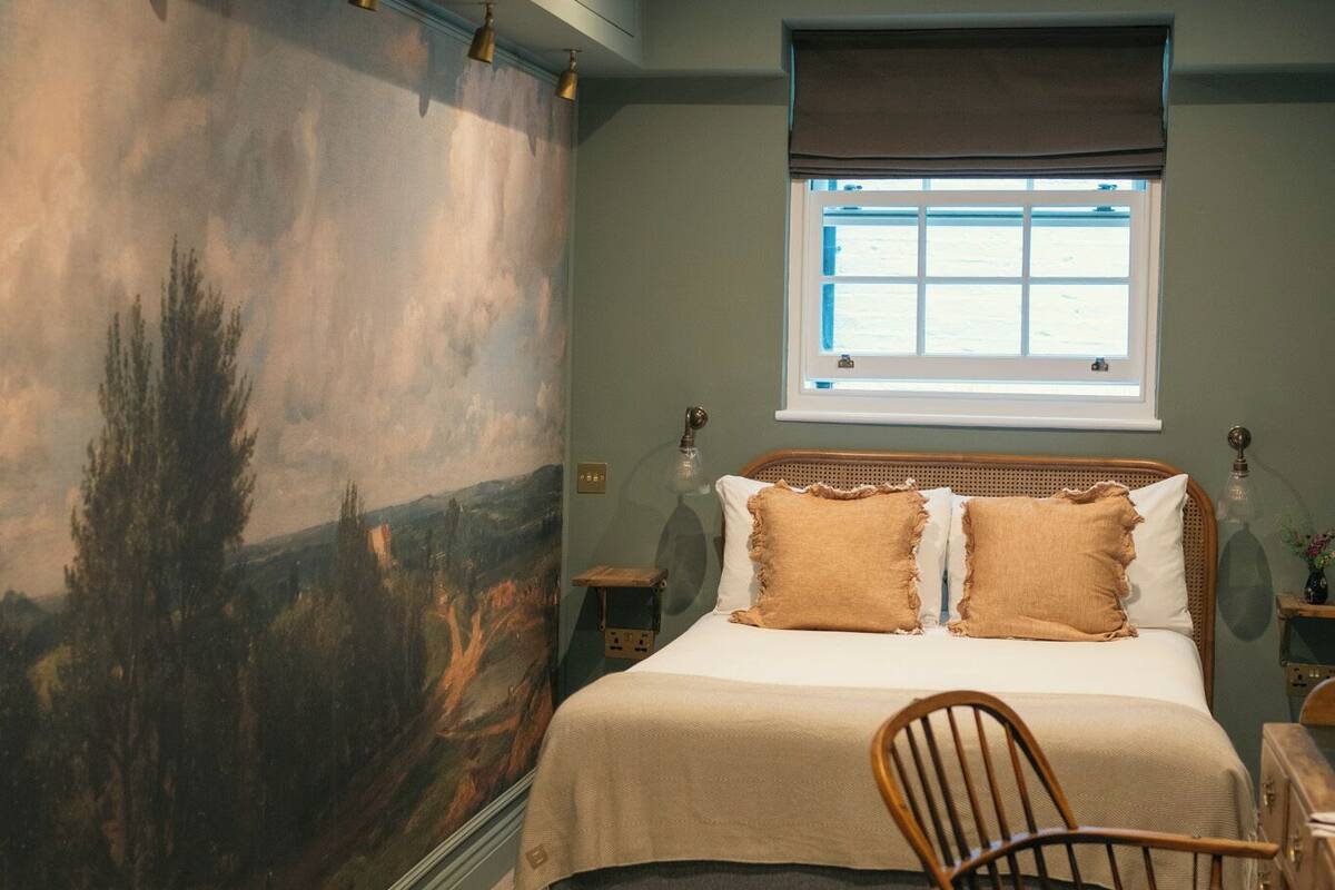 A bedroom with a large landscape mural, a bed with two pillows, a window with blinds, and a chair.