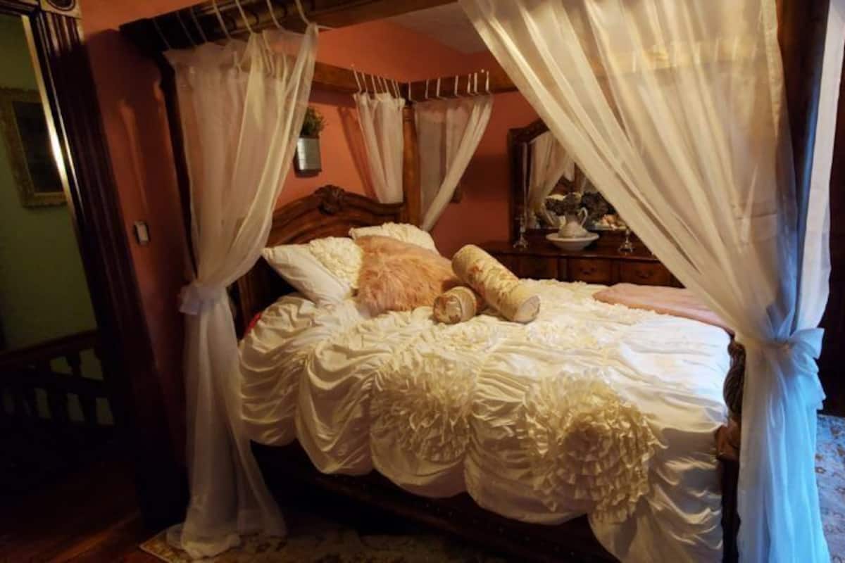 A four-poster canopy bed with white linens and pillows, a wooden headboard, and a bedside table with a mirror and decorative items.