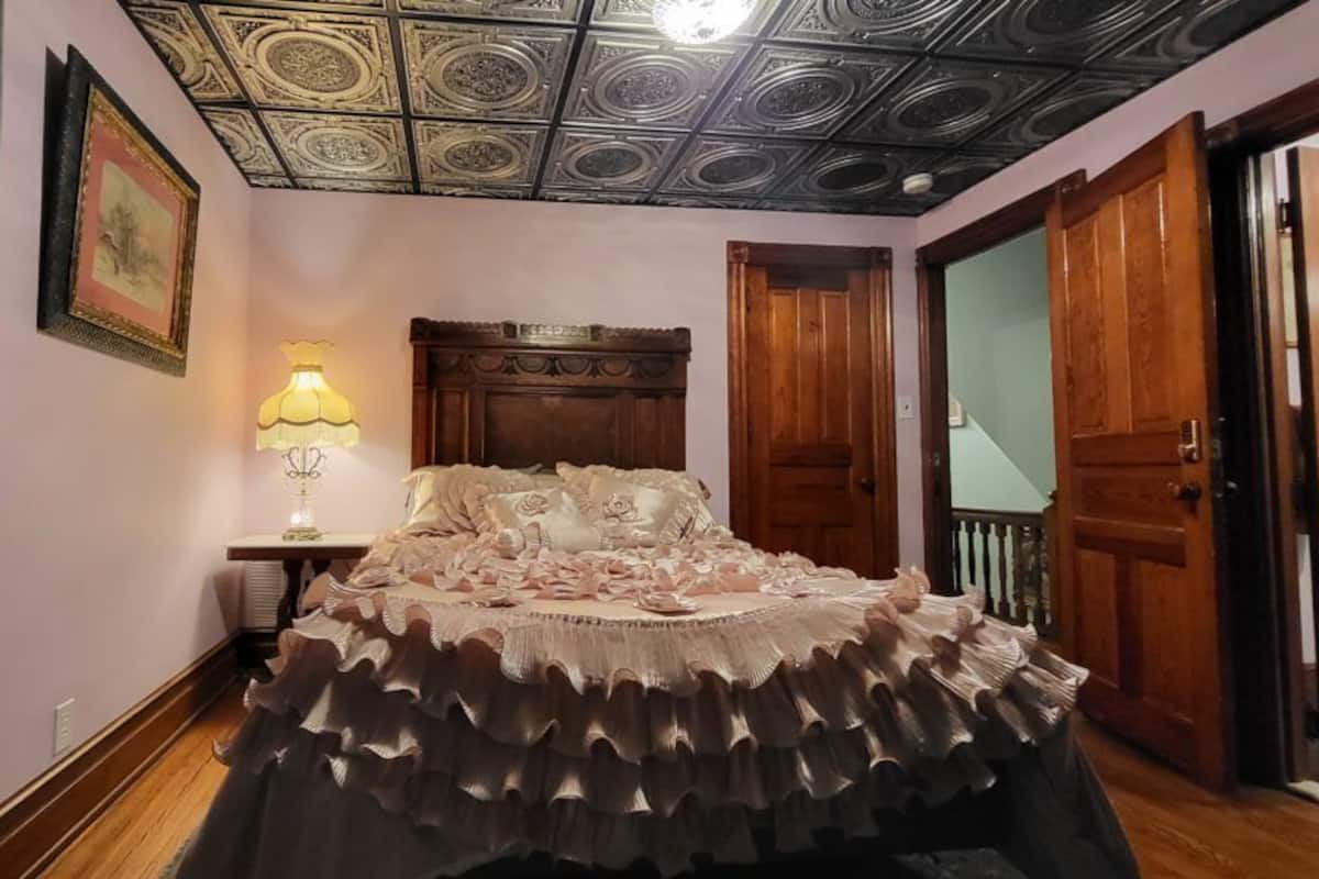 A bedroom with a ruffled bedspread, a wooden headboard, a nightstand with a lamp, and a ceiling with intricate patterns.