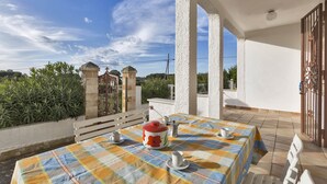 A terrace with a checkered tablecloth, a coffee pot, and cups, overlooking a garden and a gate.