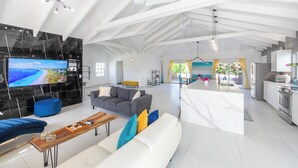 Amplia sala de estar con un televisor grande, un sofá blanco, un reposapiés azul y una cocina con armarios blancos y electrodomésticos de acero inoxidable.