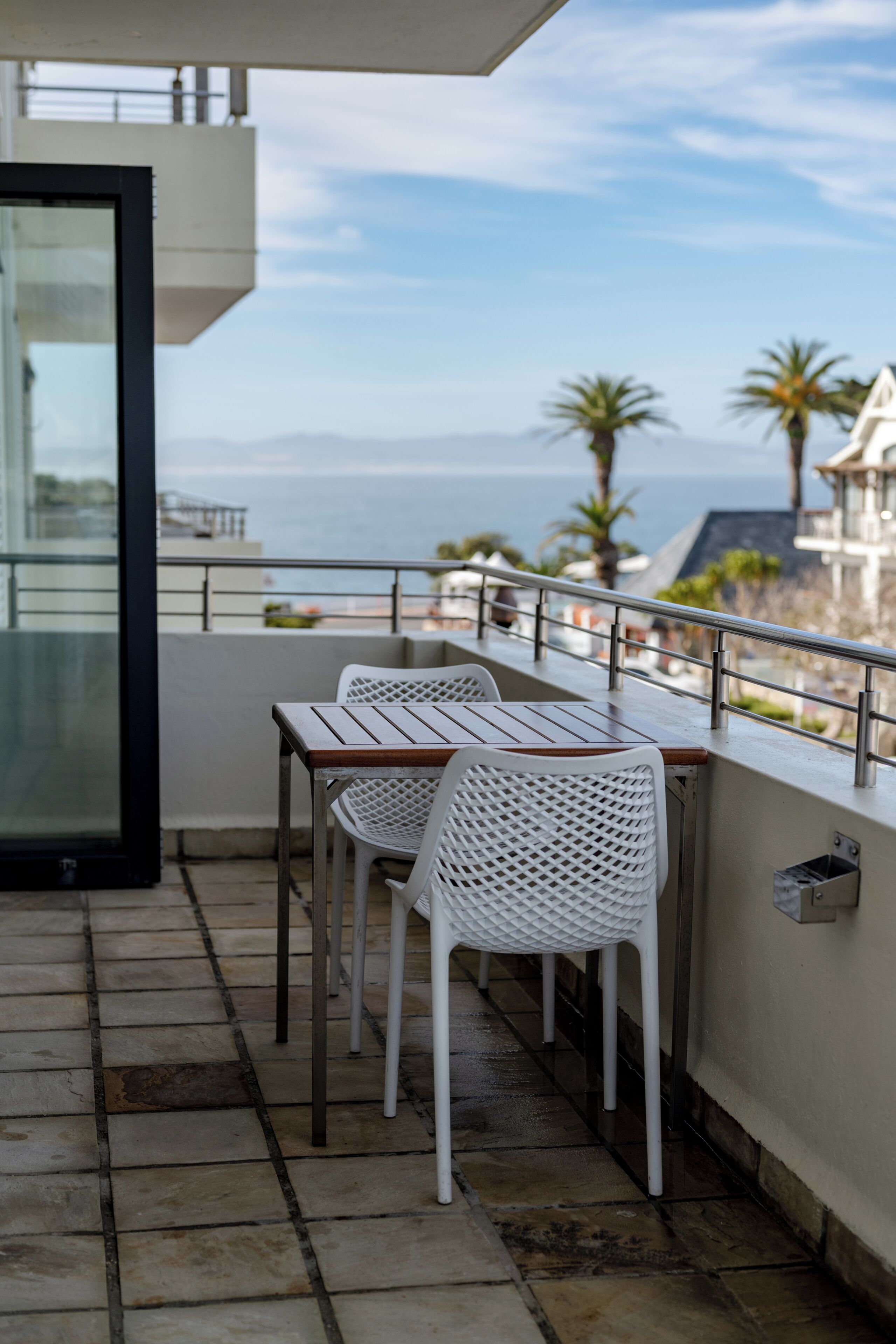 A balcony with two white chairs and a table, overlooking a coastal view with palm trees and a building in the background.