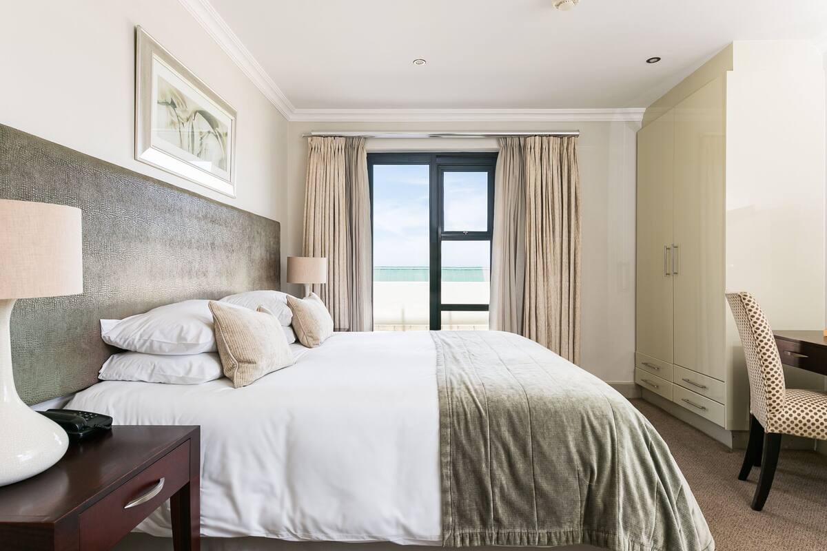 Quarto de hotel com uma cama, escrivaninha, cadeira, luminária, armário e vista para o mar.