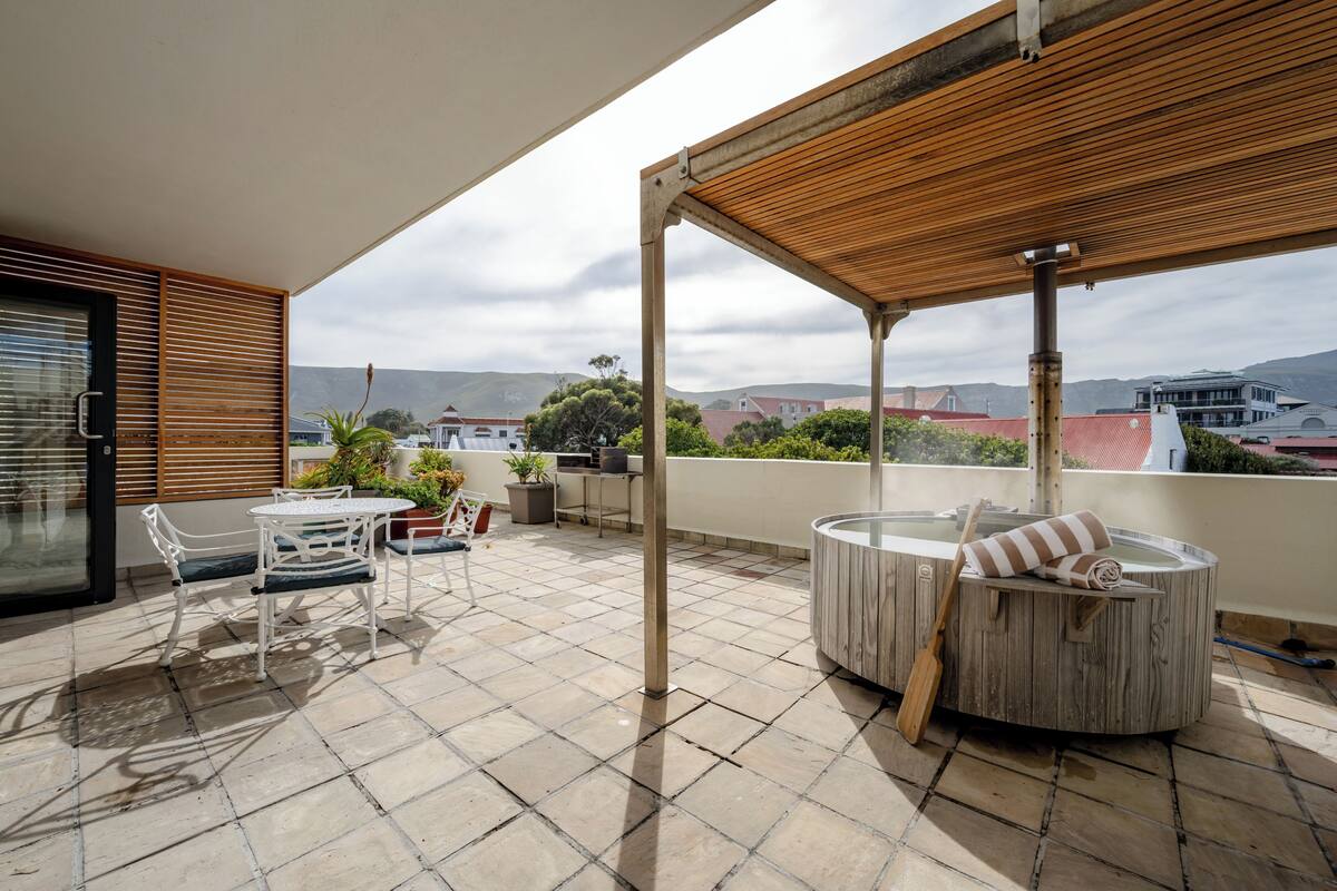 Um alpendre coberto com mesa e cadeiras de madeira, uma banheira de hidromassagem e vista para a cidade.