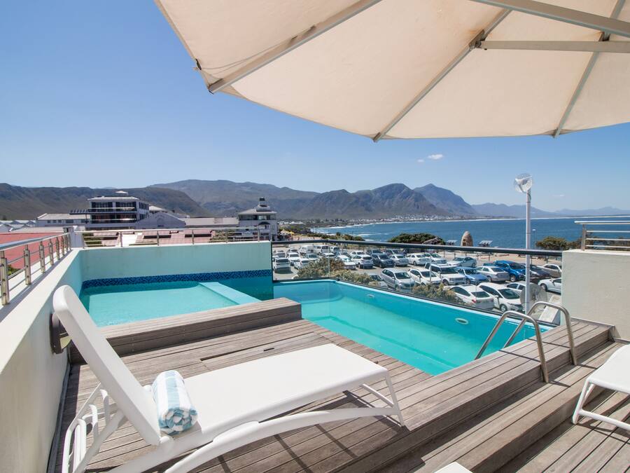 Un espace piscine aménagé sur le toit, offrant une vue sur l’océan et les montagnes, avec des chaises longues et un coin salon abrité.