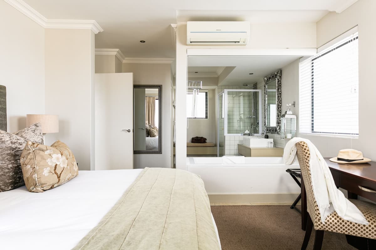 Quarto de hotel com cama, cadeira, escrivaninha, espelho, banheiro com banheira e ar condicionado.