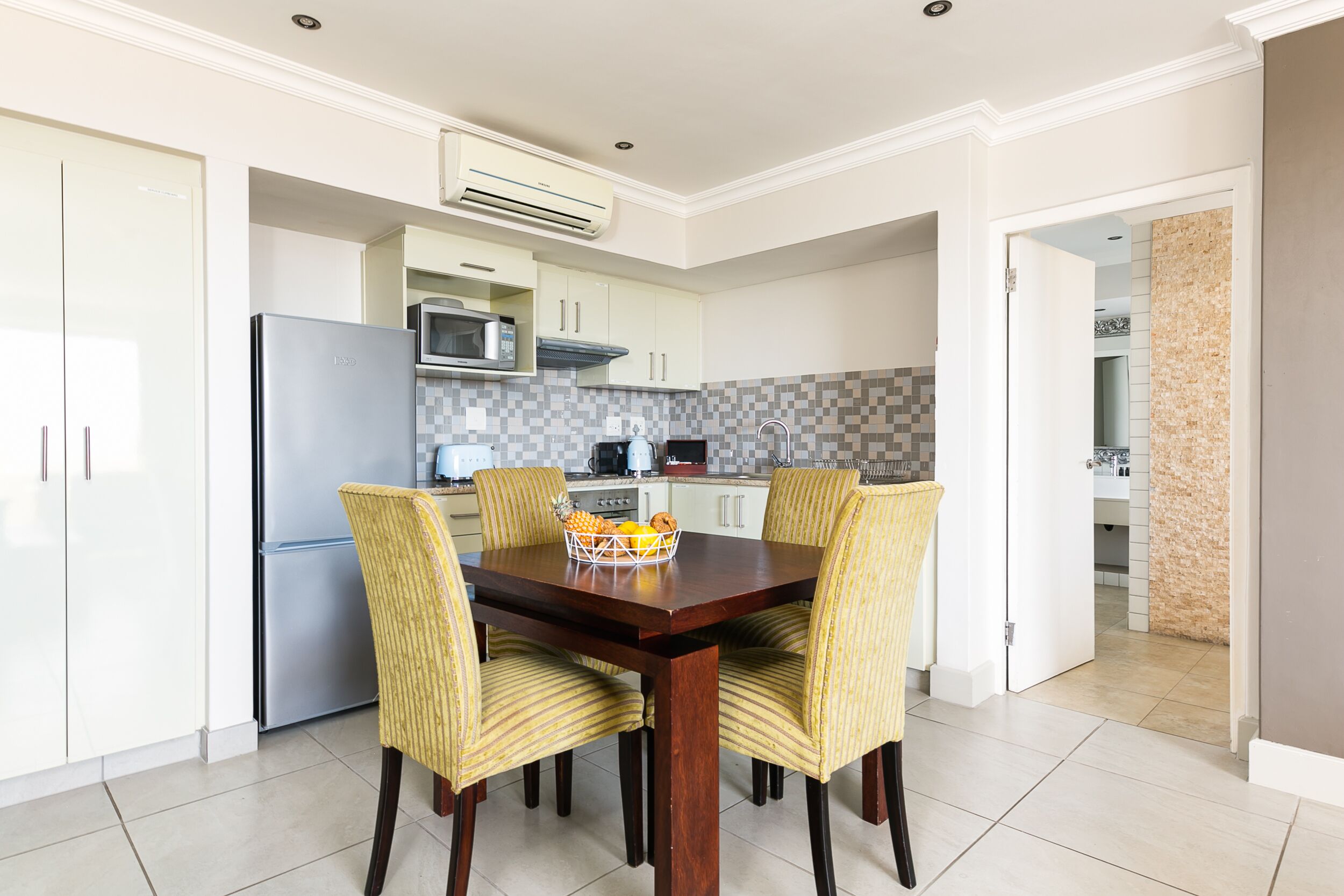 Una cocina moderna con armarios blancos, un refrigerador de acero inoxidable y un microondas. Hay una mesa de comedor con cuatro sillas a rayas y una cesta de fruta.