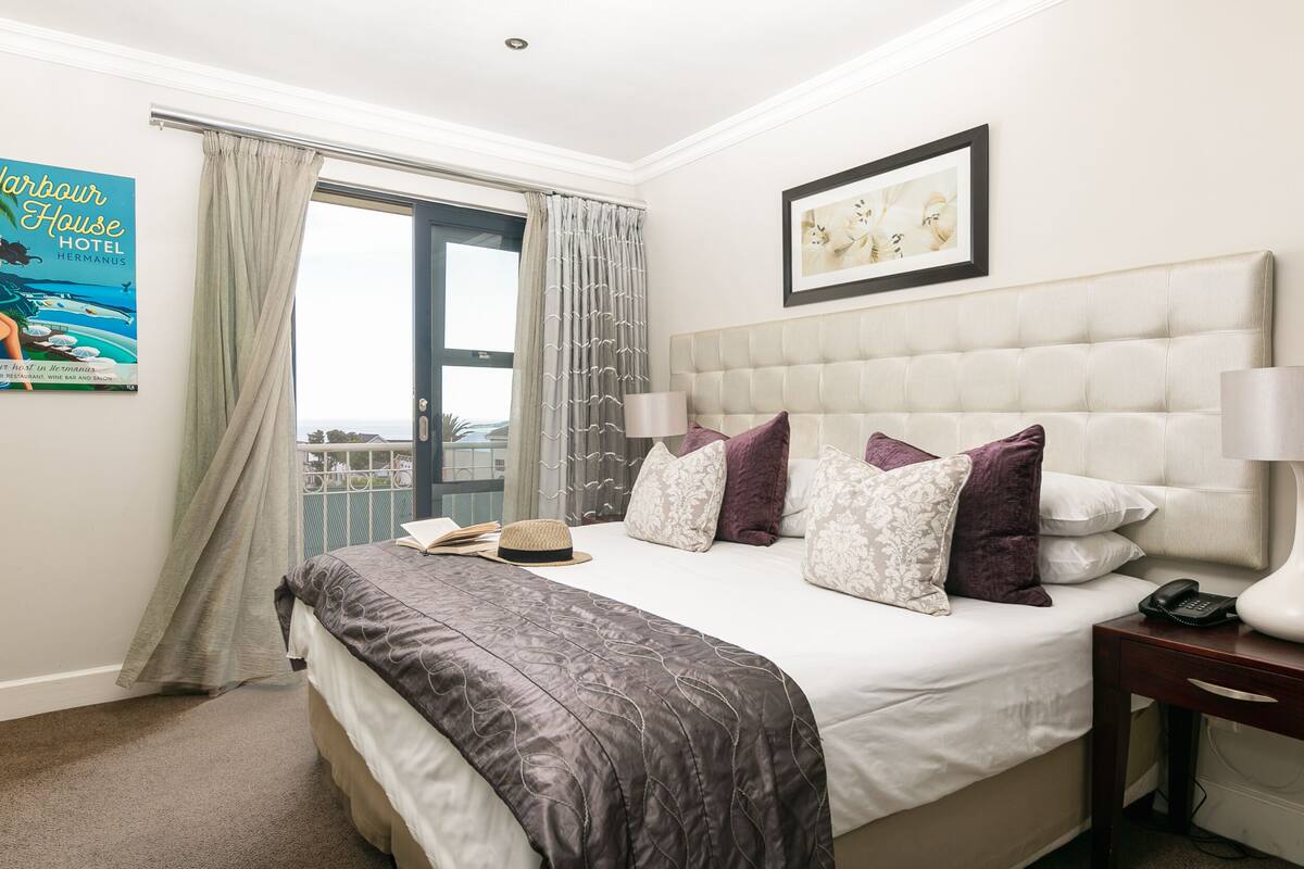 Quarto de hotel com uma cama grande, uma mesa de cabeceira, uma luminária, um quadro na parede e vista para o mar pela janela.