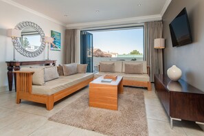A modern living room with a sofa set, a wooden coffee table, a TV, and a large window with curtains. - Harbour Square Hotel (Hermanus)