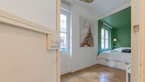 Une chambre à coucher avec un lit, une fenêtre, une lampe fixée au mur et un tableau représentant la Tour Eiffel.