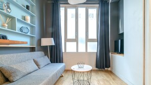 Un salon moderne avec un canapé gris, une table ronde dotée d’un pied en fil de fer, un téléviseur posé sur une étagère et une fenêtre avec des rideaux.