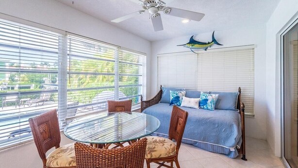 A balcony with a glass table, wicker chairs, a bed with blue bedding, and a ceiling fan. - Key Colony Beach Paradise (Key Colony Beach)
