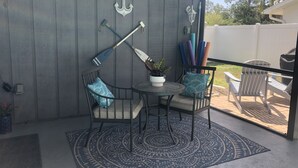 Un patio couvert comprenant deux chaises en métal, une petite table ronde, un tapis décoratif et une plante en pot.
