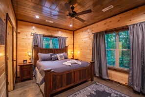 Un dormitorio de cabaña de madera con una cama, mesitas de noche, un ventilador de techo y ventanas amplias con cortinas.