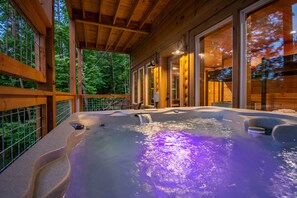 Un jacuzzi con cascada, rodeado por una terraza de madera y amplios ventanales con vistas a un bosque.