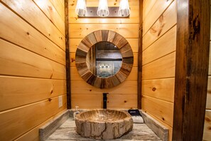 Baño de cabaña de madera con espejo circular, paredes de madera y un lavabo central.
