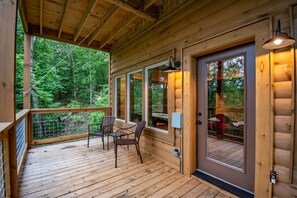 Una terraza de madera con sillas y una mesa, ventanas amplias y una puerta con vistas al bosque.