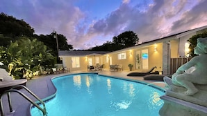 Piscina con un banco hundido, rodeada de mobiliario exterior y un edificio con varias ventanas.