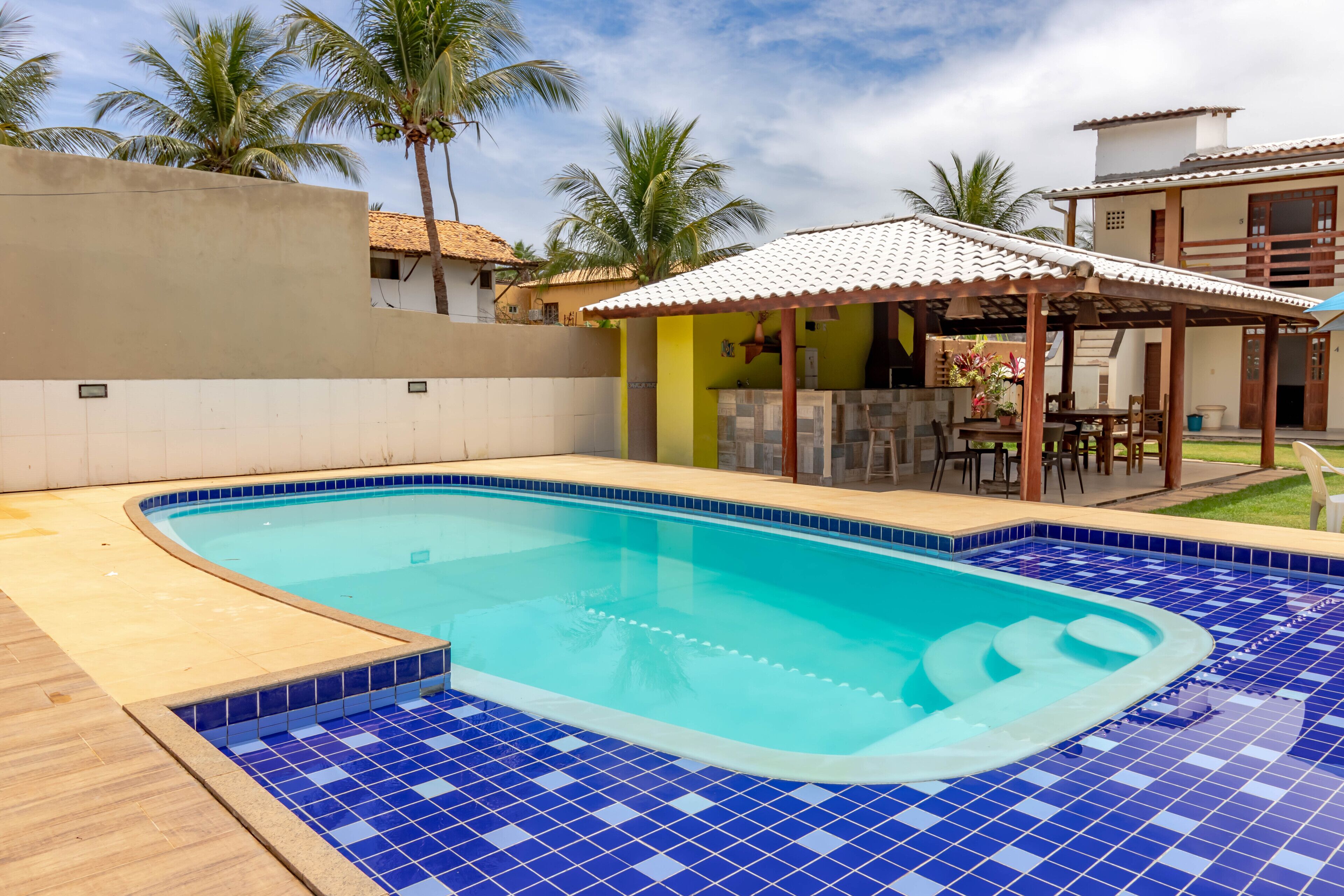 A swimming pool with blue tiles, a covered patio, and palm trees.