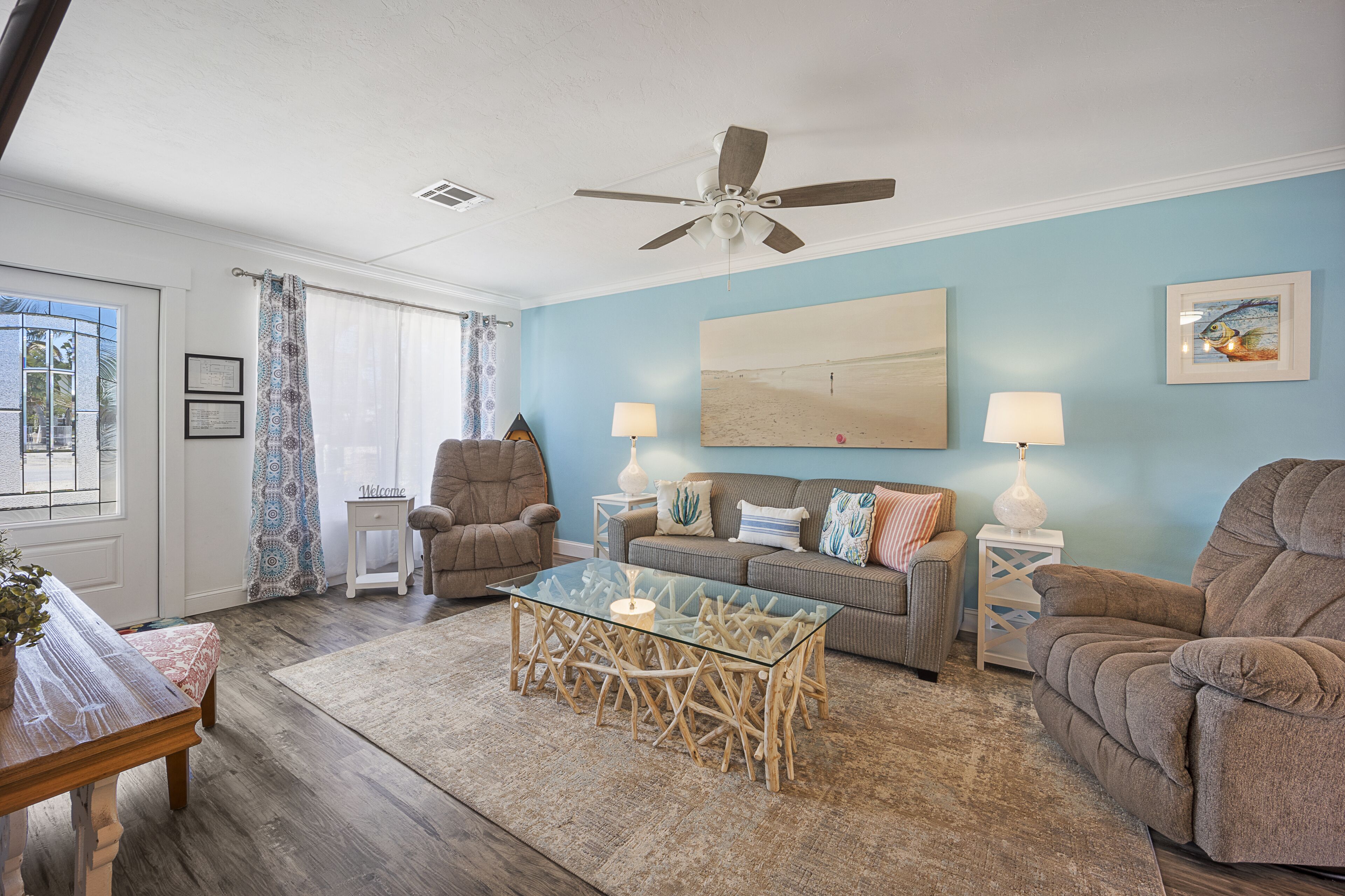 A living room with a glass coffee table, two sofas, a chair, a ceiling fan, and a large painting on the wall.