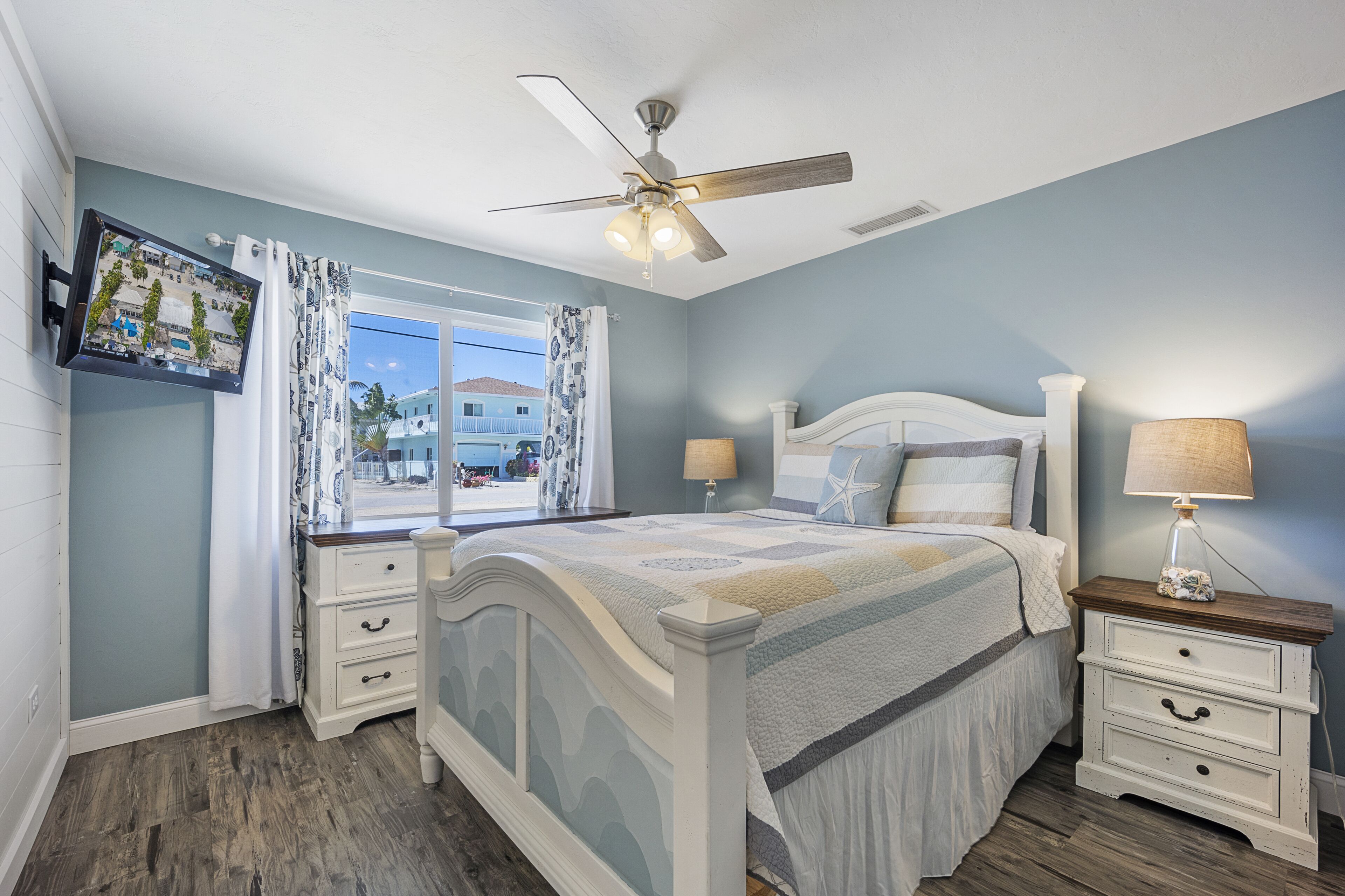 A bedroom with a bed, nightstands, a ceiling fan, a window with curtains, and a view of the outside.