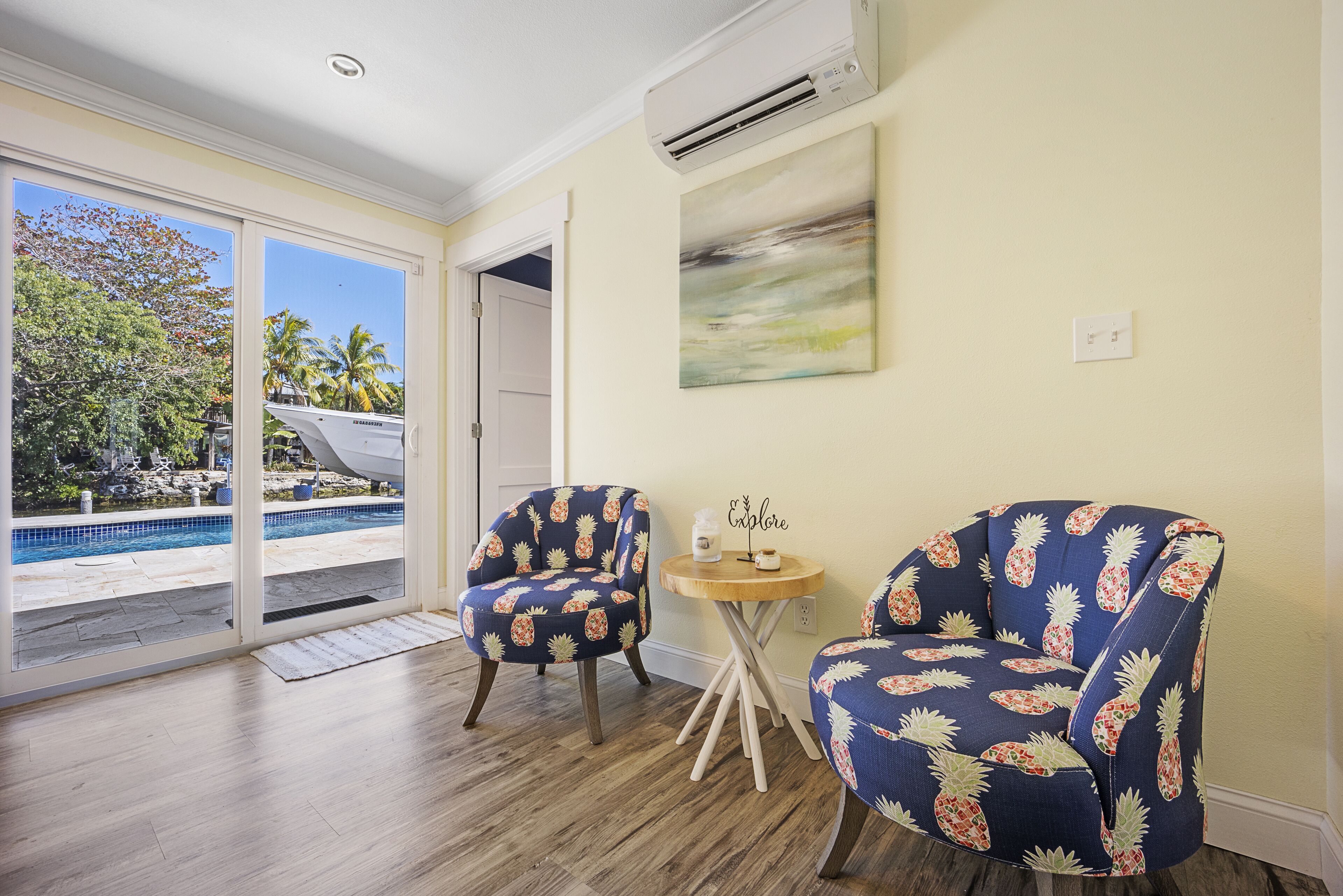 A room with a wooden floor, two chairs with a pineapple pattern, a small round table, and a painting on the wall.