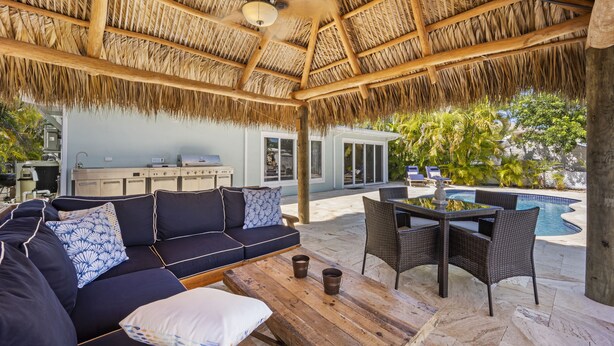A covered outdoor seating area with a thatched roof, a pool, and a dining table with chairs. - Waterfront Oasis w/ Pool, Tiki Lounge, Dock & BBQ (Marathon)