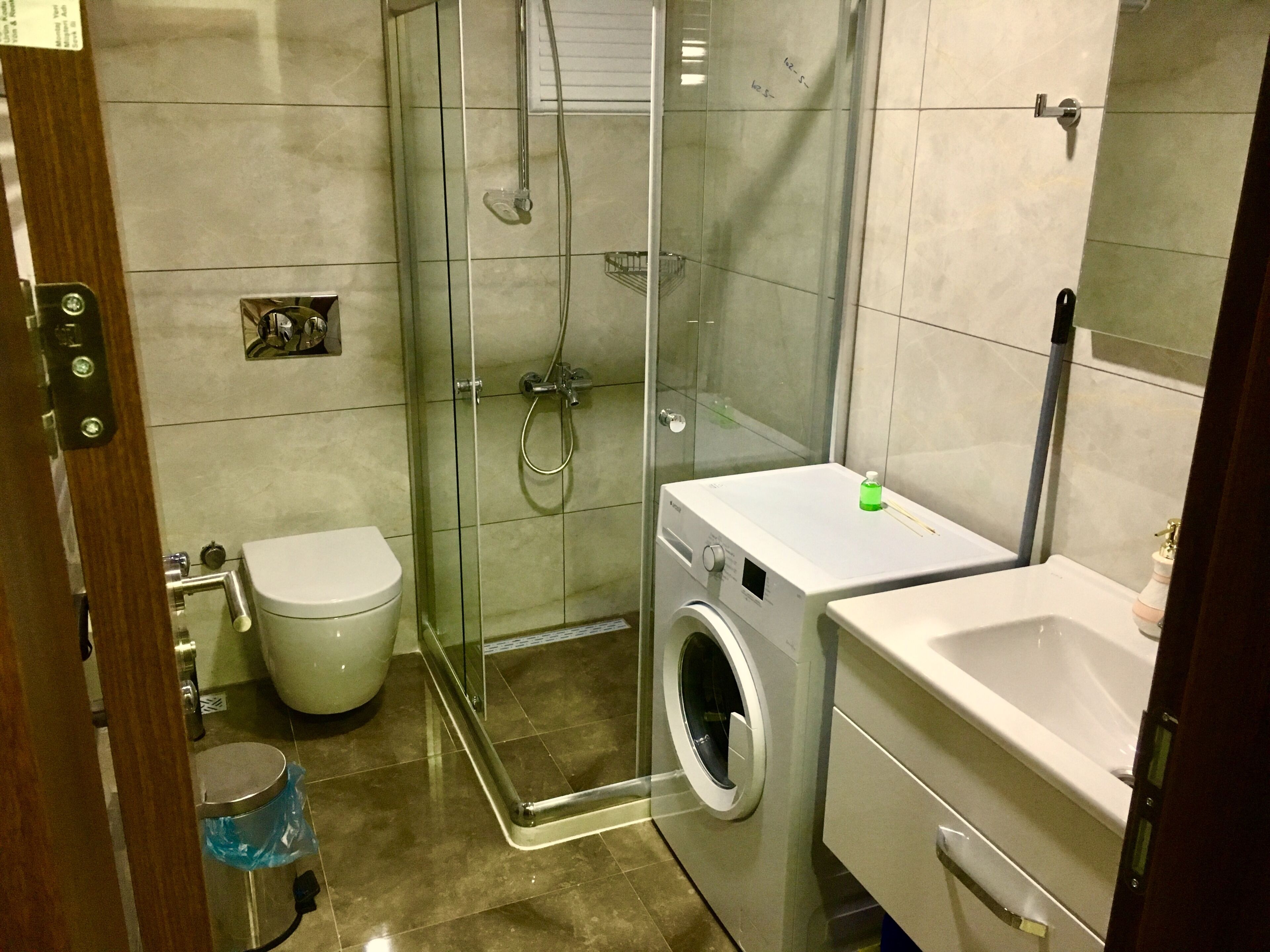 A compact bathroom with a glass-enclosed shower, a toilet, a washing machine, and a sink.