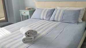 A neatly made bed with a striped bedspread and pillows, a small bedside table with a lamp, and a window with curtains. - Cozy newly built cottage, within walking distance of the center. (VILLASIMIUS)