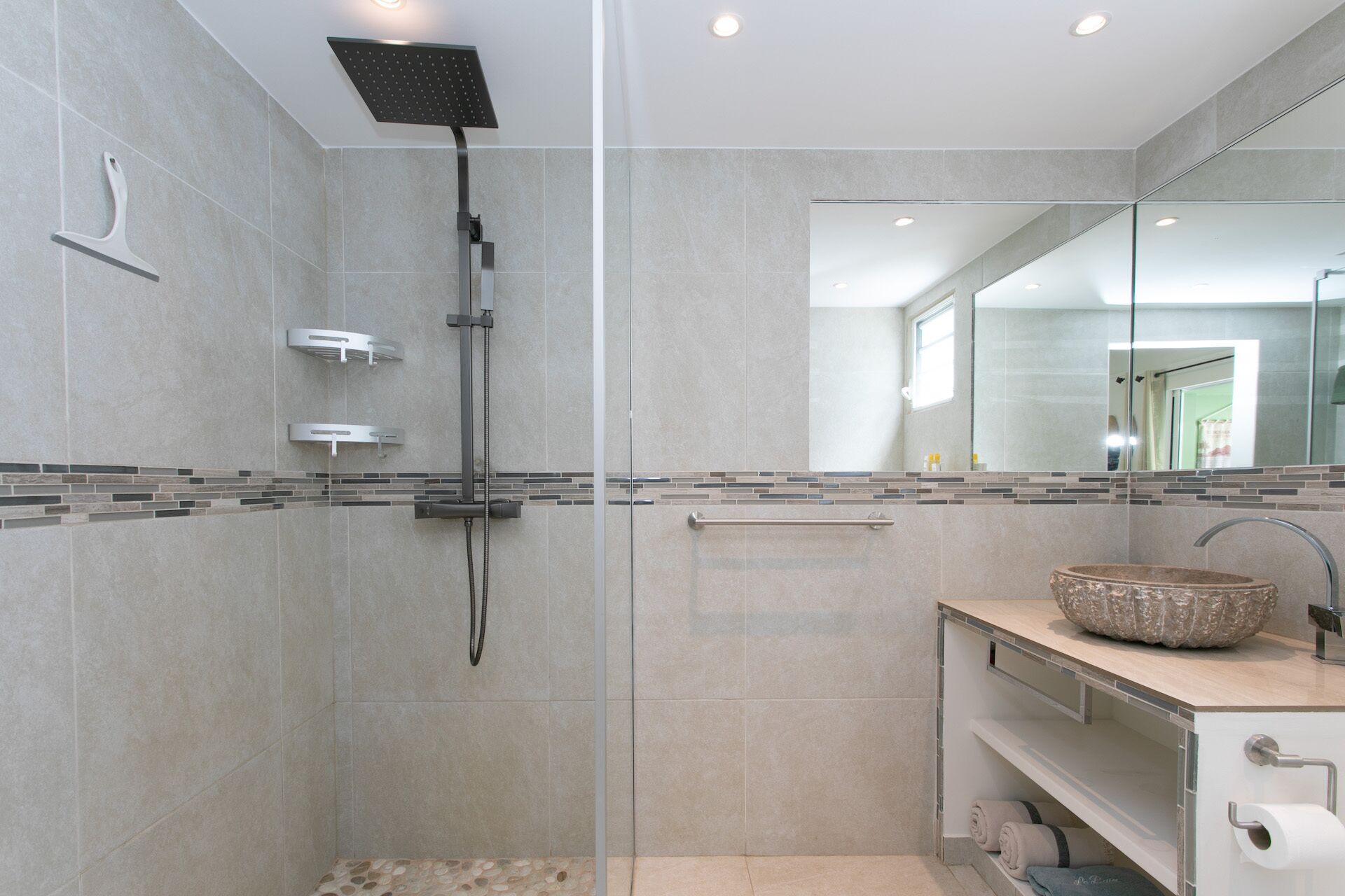 Un baño moderno con ducha envuelta en vidrio, tocador con lavamanos de mosaico y un espejo grande.