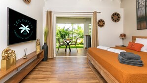 A hotel room with a bed, a TV, a wooden floor, a balcony with a view of greenery, and a dining area with chairs and a table. - Les Galets Sxm (Orient Bay)