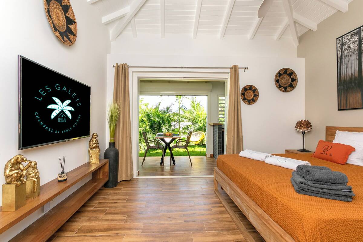 A hotel room with a bed, a TV, a wooden floor, a balcony with a view of greenery, and a dining area with chairs and a table.