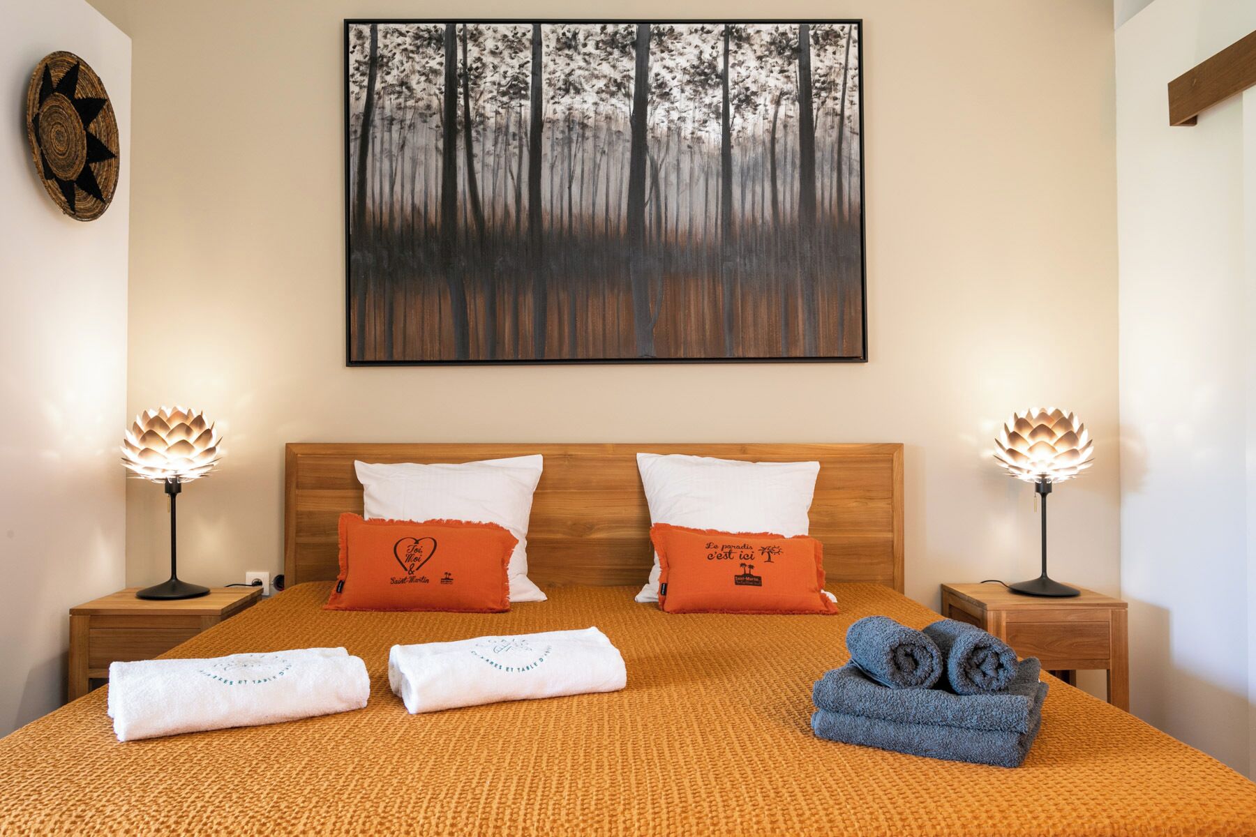 A neatly made bed with an orange blanket, two white pillows, and a red heart-shaped pillow. There are two bedside lamps with a lotus design, a framed forest painting on the wall, and a wooden headboard.
