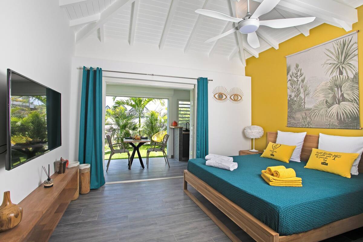 A bedroom with a bed, a TV, a ceiling fan, and a view of the outdoors.