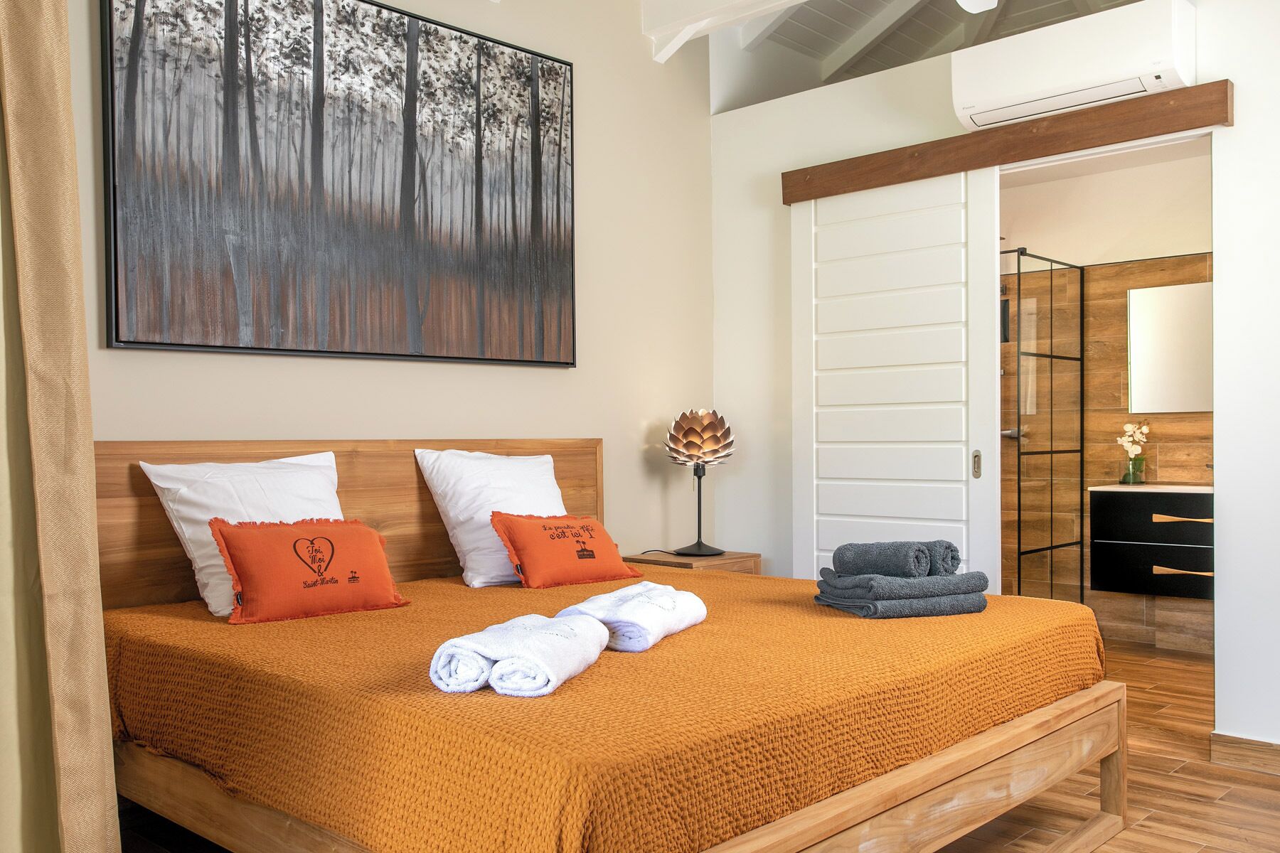 A hotel room with a wooden bed, two pillows, a bedside lamp, a towel, and a painting on the wall.