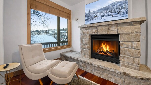 Un salon chaleureux avec une cheminée en pierre, un confortable canapé beige, un téléviseur fixé au-dessus de la cheminée et une fenêtre donnant sur un paysage enneigé.