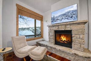 A cozy living room with a stone fireplace, a comfortable beige sofa, a television mounted above the fireplace, and a window with a snowy view. - Mountain Luxury with Front Row Access to Adventure 502B (Park City)